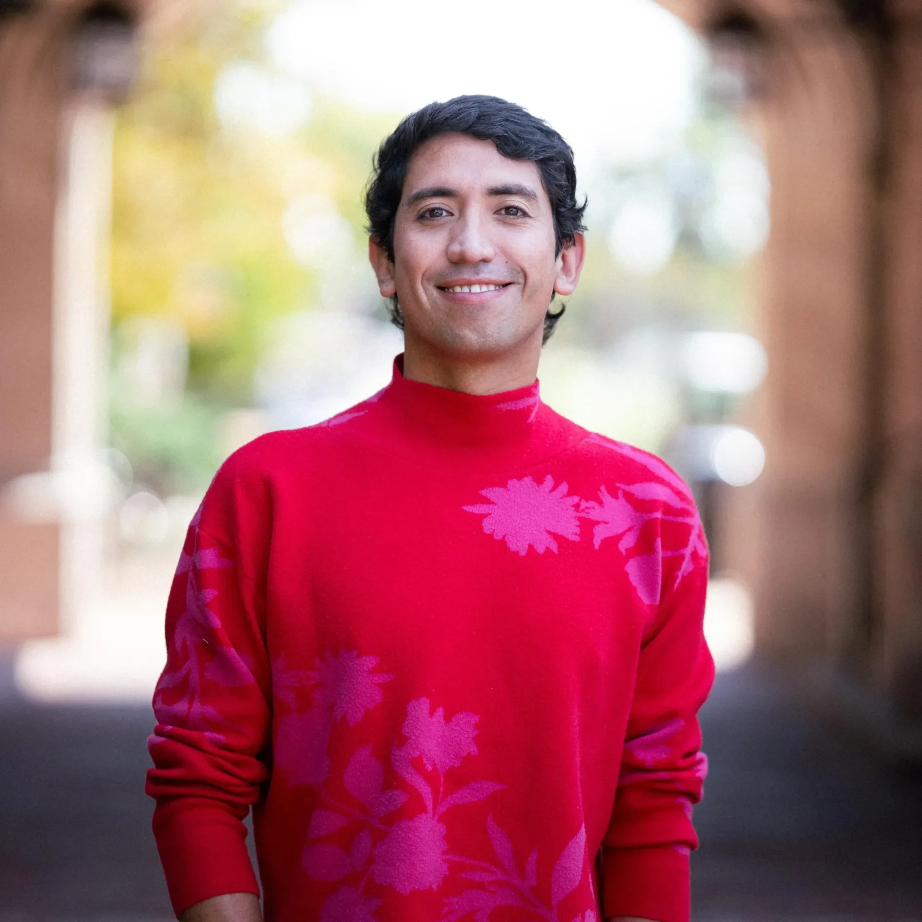 Smiling man wearing a bright red sweater with pink floral patterns, standing outdoors.