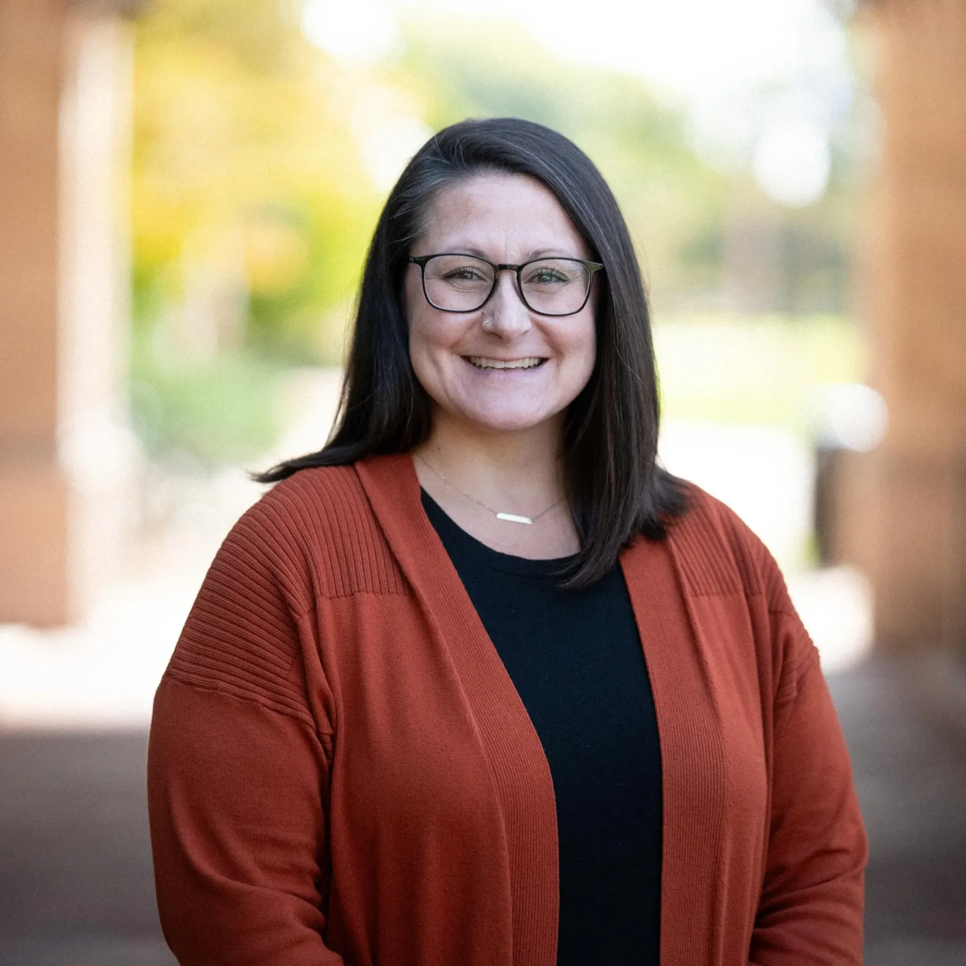 Smiling woman with glasses wearing a rust-colored cardigan and black top, standing outdoors.
