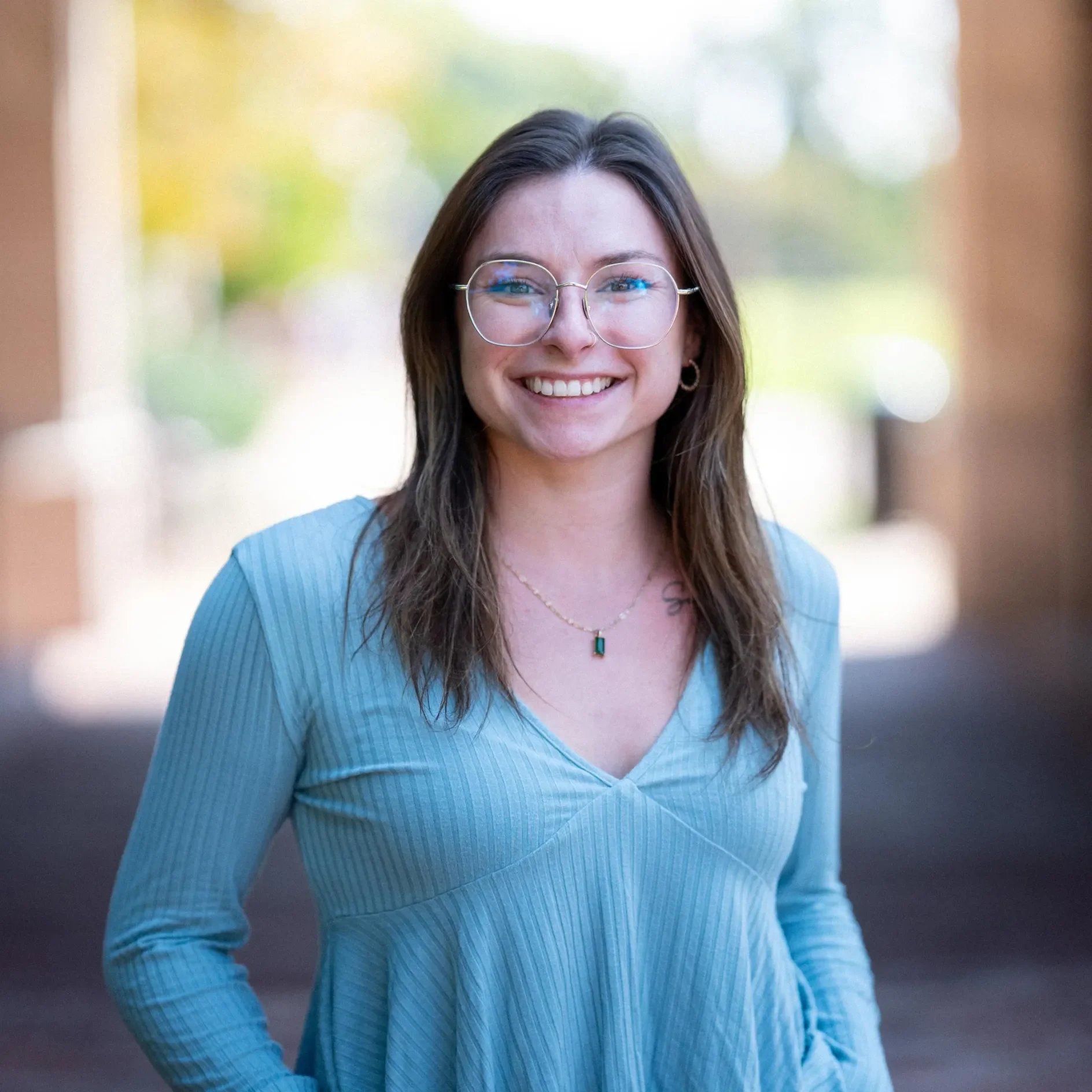 A smiling woman with long brown hair wearing round glasses and a light blue ribbed V-neck top stands outdoors in a softly blurred walkway with natural light. She is wearing a necklace with a green pendant and has her hands in her pockets.